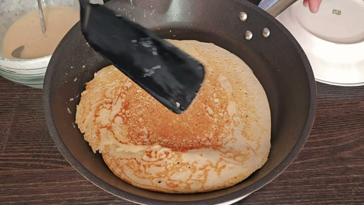 Пышный дрожжевой блин с золотистой ажурной корочкой переворачивают лопаткой на сковороде