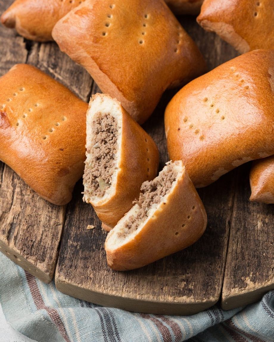 Квадратные пирожки с мясом на деревянной доске, два разломлены и видна мясная начинка